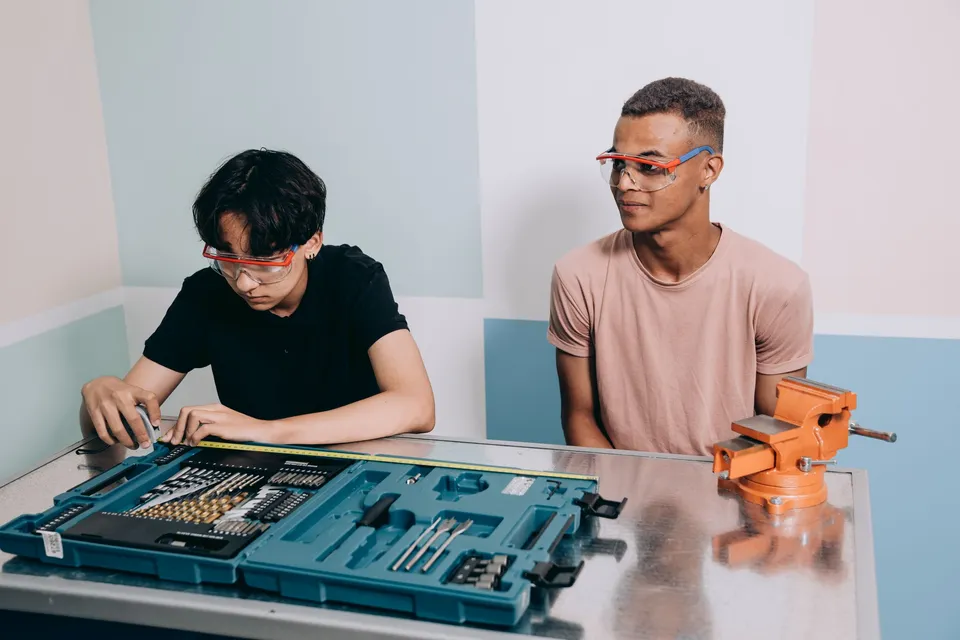 Two young men in safety glasses at a workbench with a tool kit