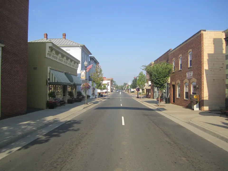 Center Street in Old Town Manassas, Virginia, looking down the main street past brick storefronts, hanging flower baskets and American flags on a clear morning